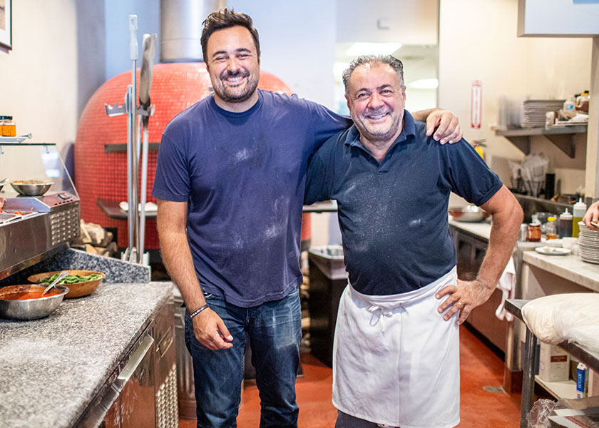 Father and son standing in the kitchen of their pizzeria.