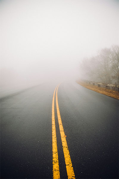A two-lane road disappears into the morning fog as it moves around a bend.
