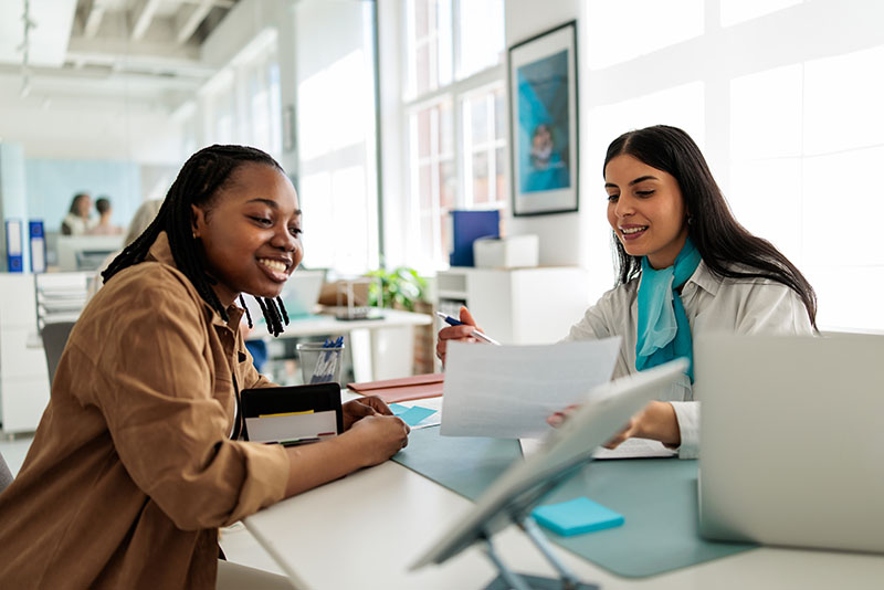 Professional woman in business attire seated at her desk in a modern bank office, discussing financial matters with a female customer.